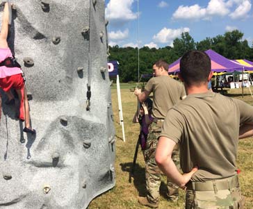 A woman climbing a rock wall with two military personnel standing next to it.