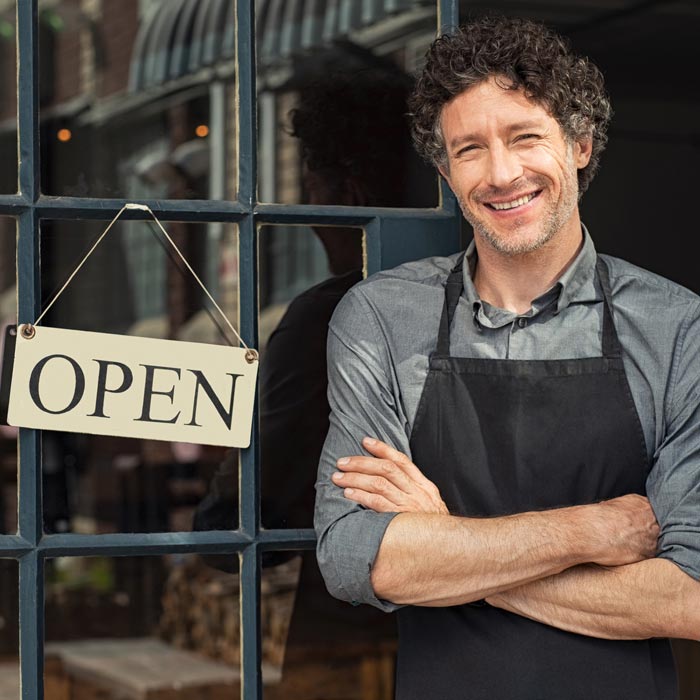 A Smiling man crossing his arms, standing in front of his restaurant next to the open sign.